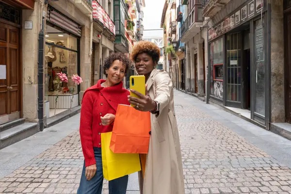 İki mutlu bayan arkadaş, parke taşlı bir sokakta alışveriş torbaları tutarken akıllı telefonlarıyla selfie çekiyorlar.