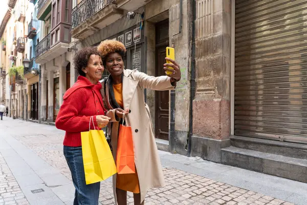 İki çok ırklı kadın ellerinde renkli poşetlerle alışveriş sonrası akıllı telefonlarıyla selfie çekiyor.