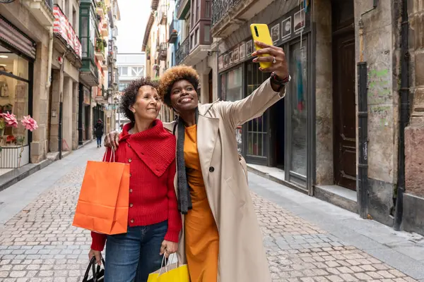 İki mutlu bayan arkadaş şehir merkezinde alışveriş torbaları taşırken akıllı telefonlarıyla selfie çekiyorlar.