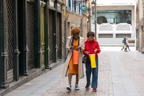 İki kadın, renkli alışveriş çantalarının içine bakıp gülümseyen yaya alışveriş caddesinde yürüyorlar.