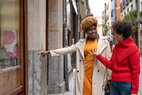 Şehir caddesinde çene çalarken iki bayan arkadaş yürüyor ve vitrinlere bakıyor. Biri dükkanın camına bakıyor.