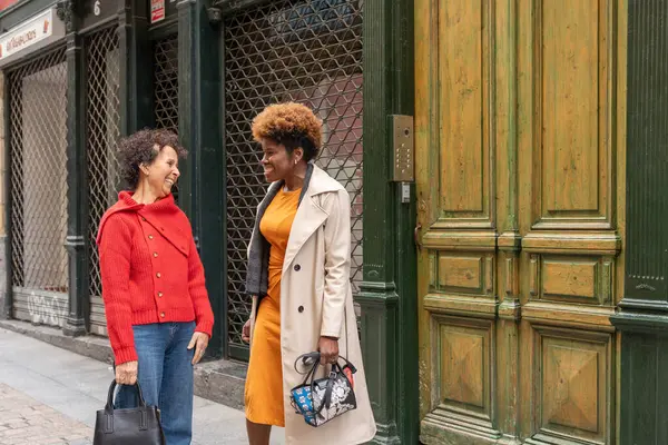 Biri kırmızı kazaklı, diğeri bej renkli trençkotlu iki kadın, şehrin kaldırımında durup gülümsüyor ve yıpranmış yeşil bir kapının yanında konuşuyorlar.