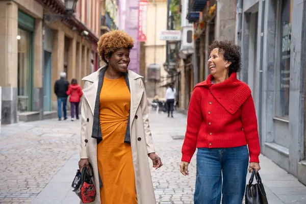 Kalabalık bir caddede yürüyen ve sohbet eden iki neşeli kadın alışveriş yaparken ve şehir manzarasını keşfederken birlikte vakit geçiriyorlar.