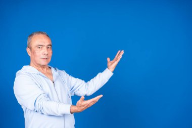 Sıradan kıyafetler giyen yaşlı adam mavi arka planda bir şey sunan elleriyle el kol hareketi yapıyor.