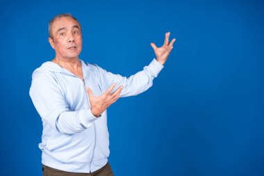 Açık ellerle işaret eden açık renk ceketli olgun bir adam, açık mavi bir arkaplan üzerine kurulmuş, iletişim ve duygu iletiyor.