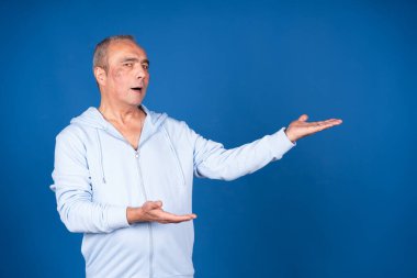 Normal giyinen olgun bir adam mavi arka planda elleri olan bir şeyi gösteriyor ve açıklıyor.