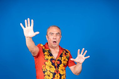 Hawaii gömleği giyen kıdemli bir adam korku ve şaşkınlık jesti içinde paniğe ve endişeye işaret eden canlı bir mavi arka plana el kaldırıyor.