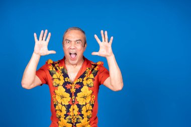 Renkli gömlek giyen etkileyici olgun bir adam elleri ile hareket ediyor ve parlak mavi bir arka planda şaşkınlık ve mutluluk gösteriyor.