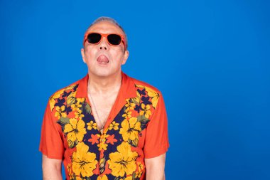 Güneş gözlüğü takan ve Hawaii gömleği giyen neşeli bir son sınıf öğrencisinin neşeli mavi arka planına dilini uzattığı bir fotoğraf.