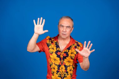 Havai gömleği giyen kıdemli adam el kaldırıyor, dur işareti veriyor, reddediyor ya da canlı mavi arka planda savunma yapıyor.