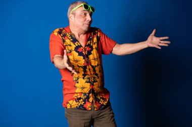 Renkli bir tişört ve güneş gözlüğü takan son sınıf öğrencisi mavi arka planda komik bir ifade ve açık kollu karışık bir jest yapıyor.