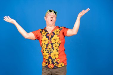 Hawaii gömleği ve güneş gözlüğü takan, kollarını mavi bir arka plana karşı açık omuz silken, şaşırmış, olgun bir adamın stüdyo fotoğrafı.