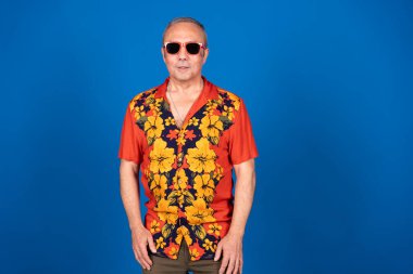 Canlı bir Hawaii gömleği ve şık bir güneş gözlüğü takan olgun bir adam mavi bir stüdyo arkaplanına gülümserken tasasız bir tatilin tadını çıkarıyor.