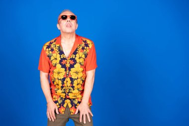 Canlı Hawaii gömleği ve güneş gözlüğü takan yaşlı bir adam, şakacı bir şekilde dilini dışarı çıkarıyor, kaygısız bir tutum sergiliyor.