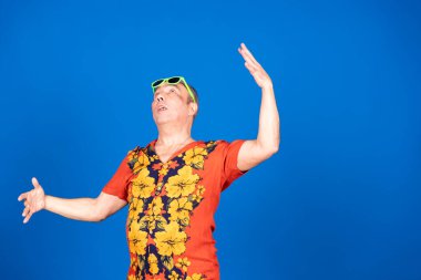 Hawaii tişörtü ve güneş gözlüğü takan orta yaşlı bir adam mavi arka planda el kaldırıyor.