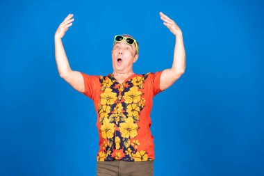 Havai gömleği ve güneş gözlüğü takan olgun bir adam mavi arka planda şaşkınlıkla kollarını kaldırıyor.