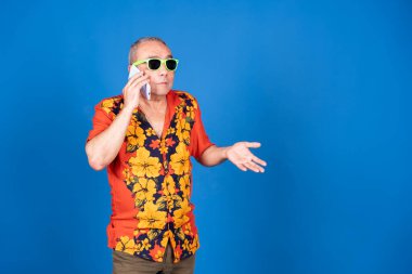 Orta yaşlı, güneş gözlüğü takan ve Hawaii gömleği giyen bir adam telefonda konuşurken mavi arka planla konuşuyor.