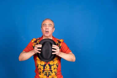 Hawaii gömleği giyen orta yaşlı sihirbaz elinde siyah şapkayla mavi arka planda illüzyon yapıyor.