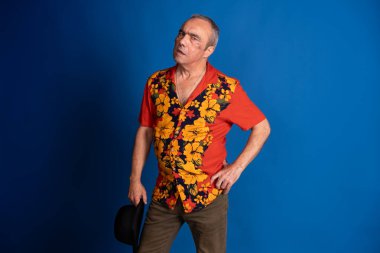 Olgun bir adam renkli bir Hawaii gömleği giyiyor ve elinde bir şapka tutuyor. Stüdyoda canlı mavi bir arka plana karşı güvenle poz veriyor.