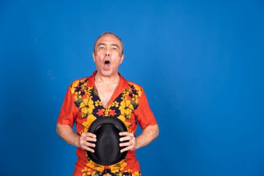 Orta yaşlı bir adam canlı bir Hawaii gömleği giyiyor, elinde bir şapka tutuyor ve parlak mavi bir arka plana karşı sürpriz bir yüz ifadesi sergiliyor.