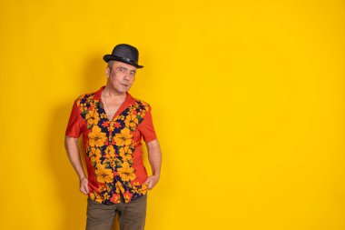 Çiçekli gömlek ve şapka giyen orta yaşlı bir adam, canlı sarı bir arkaplan karşısında duruyor, rahat ve kendinden emin bir tavır sergiliyor.