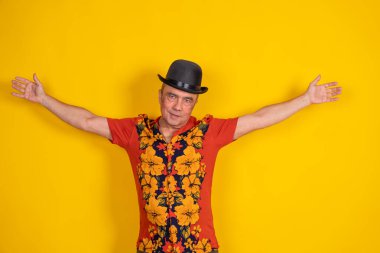 Havai gömleği ve melon şapka giyen kıdemli bir adam kollarını parlak sarı bir arka planda açıyor.