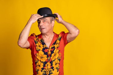 Renkli Hawaii gömlekli olgun bir adamın, siyah melon şapkasını canlı sarı bir arka plana ayarlarken çekilmiş bir fotoğrafı.