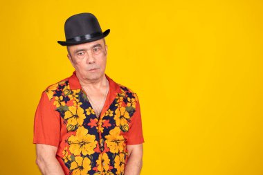 Havai gömlekli ve melon şapkalı, parlak sarı bir fonda poz veren olgun bir adamın stüdyo fotoğrafı.