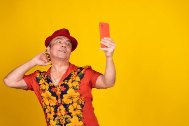 Havai gömleği ve kırmızı şapkalı son sınıf öğrencisi sarı arka planda akıllı telefonuyla selfie çekiyor.