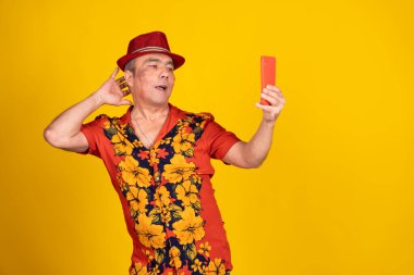 Havai gömleği ve kırmızı şapkalı etkileyici olgun bir adam sarı arka planda akıllı telefonuyla video görüşmesi yapıyor.