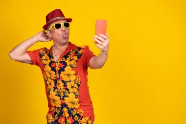 Şapkalı ve güneş gözlüklü, neşeli, olgun bir adam. Dilinden selfie çekiyor. Canlı sarı bir arka planda izole edilmiş.
