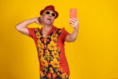 Tropikal tişörtlü ve şapkalı tutkulu orta yaşlı bir adam selfie çekiyor, parlak renklerle ve neşeli pozlarla tatil titreşimleri yayıyor.