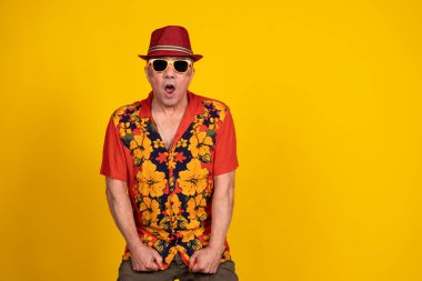 Kıdemli adam canlı bir Hawaii gömleği ve şık bir güneş gözlüğü takıyor. Parlak sarı bir arka plana karşı şaşkınlığını dile getiriyor.