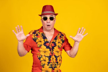 Hawaii gömleği, güneş gözlüğü ve şapka takan kıdemli bir adam sarı arka planda açık ellerini göstermesine şaşırmış görünüyor.