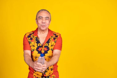Kıdemli adam canlı bir Hawaii gömleği giyiyor, parlak sarı bir arka plana karşı ferahlatıcı bir şişe su sunuyor,