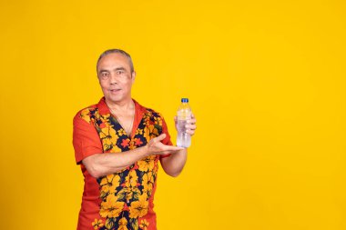 Hawaii gömleği giyen kıdemli bir adam sarı arka planda bir gülümseme olan bir şişe su sunuyor.