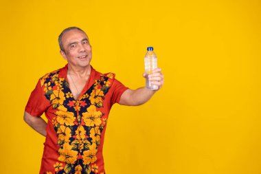 Havai gömleği giyen gülümseyen olgun bir adam canlı sarı arka planda bir şişe su sunuyor.