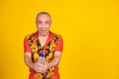 Hawai gömleği giyen etkileyici olgun bir adam sarı arka planda komik bir ifadeyle bir şişe su sunuyor.