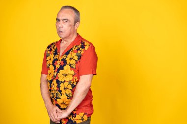 Hawaii gömleği giyen kıdemli bir adam canlı sarı bir arka planda şüpheli bir ifadeyle poz veriyor.