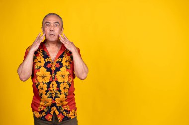 Hawaii gömleği giyen üst düzey bir adam şok ve şaşkınlık içinde, sarı arka planda elleri olan bir yüze dokunuyor.