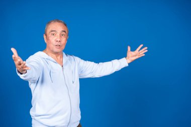 Açık renk elbiseli yaşlı bir adam kollarını mavi bir zemine doğru enerjik bir şekilde hareket ediyor. Heyecan ve pozitiflik taşıyor.