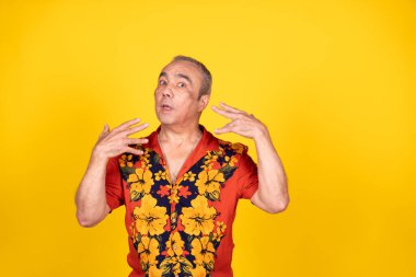 Kıdemli adam canlı bir Hawai gömleği giyiyor, parlak sarı bir arka plana el sallıyor, şaşırdığını ve merakını dile getiriyor.