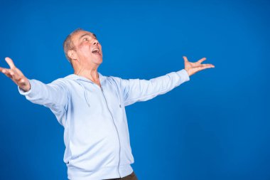Kolları açık ve yukarı bakan yaşlı bir adam, canlı mavi bir arka planda neşe ve minnettarlık ifade ediyor