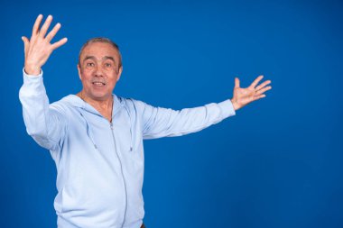 Açık renk ceketli, canlı mavi bir arka plana karşı enerjik bir şekilde el kol hareketi yapan olgun bir adam heyecan ve pozitifliği ifade ediyor.