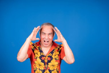Havai gömleği giyen kıdemli bir adam bağırıyor ve elleriyle başını tutuyor. Mavi arka planda çaresizlik ve stresi ifade ediyor.