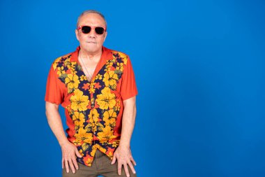 Renkli Hawaii gömleği ve güneş gözlüğü takan olgun bir adamın canlı mavi bir fon karşısında rahat ve neşeli bir yaz havasının tadını çıkardığı bir fotoğraf.