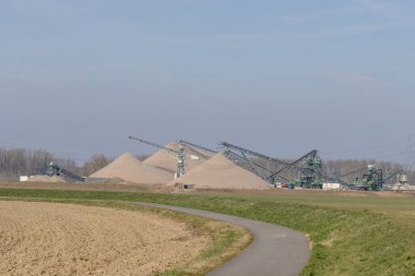 A rural industrial scene in Bobenheim-Roxheim, Germany, featuring large piles of sand, conveyor belt systems, and industrial structures under a clear blue sky. The foreground includes a curved paved path bordered by green grass and farmland, creating clipart