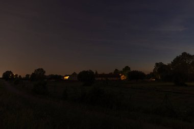 A quiet rural scene at night with softly illuminated farmhouses surrounded by trees under a starry sky. clipart