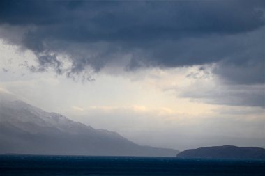 Gök gürültüsü yaklaşırken deniz manzarası. Henüz tüm gökyüzü bulutlarla kaplı değil ve uzak mesafede güneşin altındaki dağları görebilirsiniz..