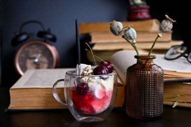 Eski kitapların yanında içinde küçük bir kaşık olan lezzetli bir tatlı ve yanında kurumuş haşhaş tohumları olan güzel bir vazo var. Arka planda ise çalışma gününün henüz bitmediğini gösteren bardaklar ve çalar saat var..